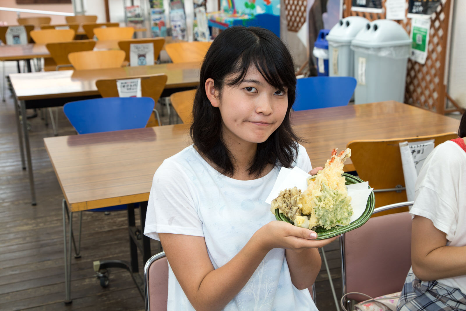 テーブルと椅子が並んだ飲食スペースで、魚や野菜の天ぷらが盛り付けられた皿を持っている女子大学生の写真