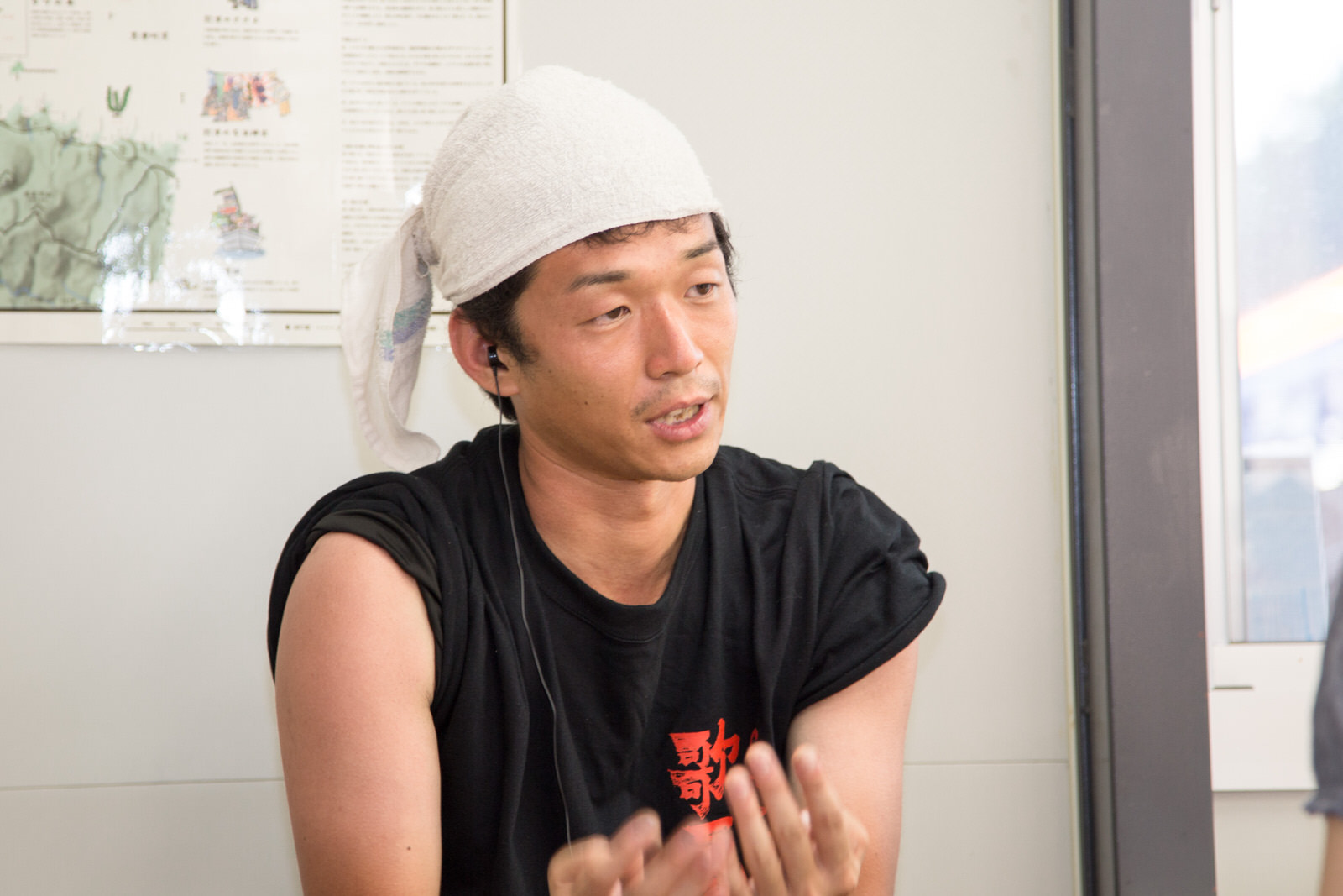 「歌夏」と書かれた黒いTシャツを着てタオルを頭に巻いた男性が、室内で真剣な表情をしながら両手を前に出して話している様子の写真