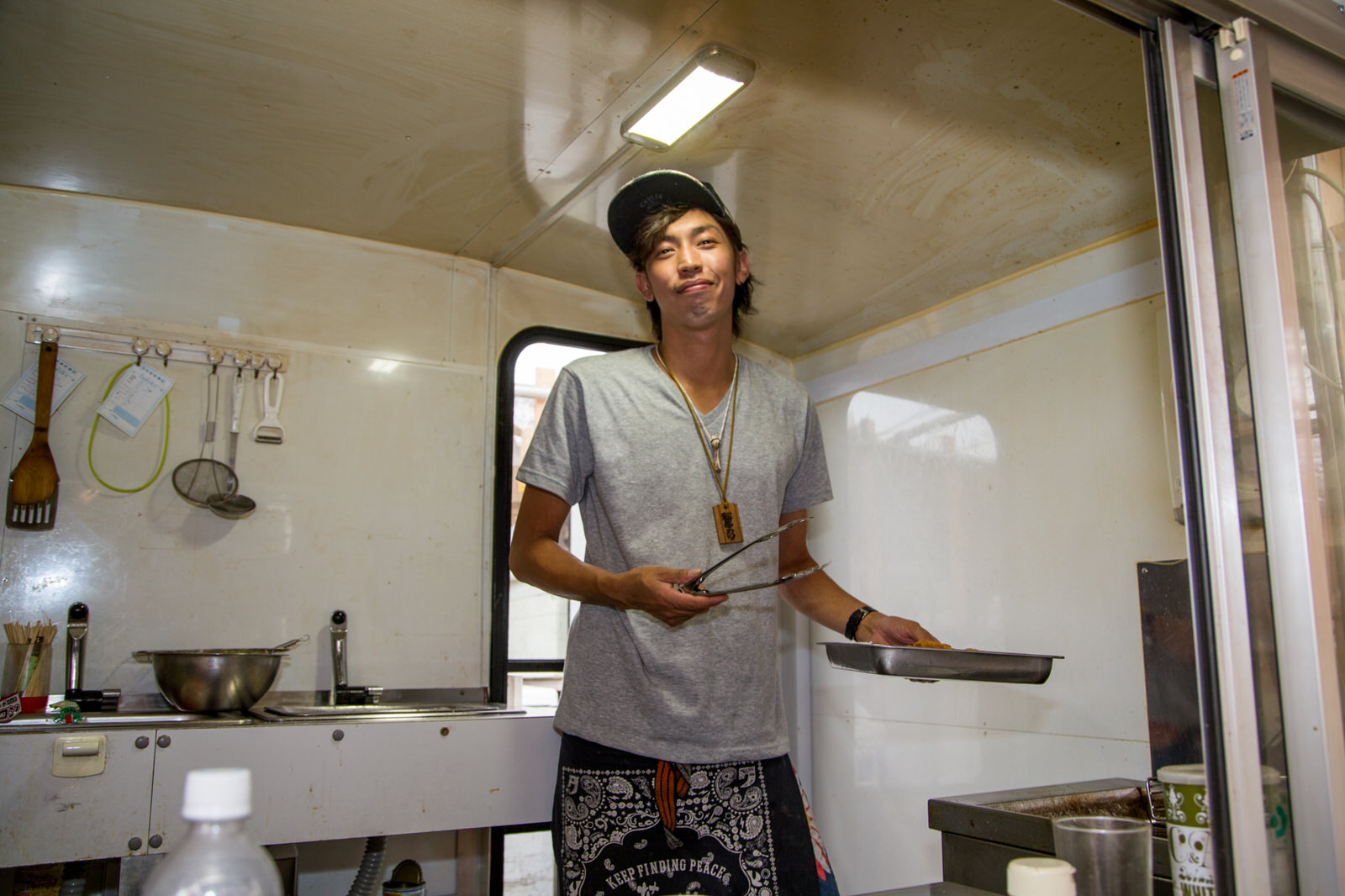 白い壁の調理スペースで、帽子と前掛けエプロンを身に着け、トングとバットを持って立っている井尻さんの写真