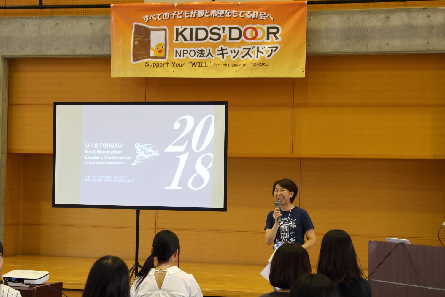 オレンジ色の横断幕に「KIDS' DOOR（キッズドア）」と書かれた横断幕が掲げられた講堂で、資料が映し出されているスクリーンの右側に立つマイクを持った女性が壇上で笑顔で話している写真