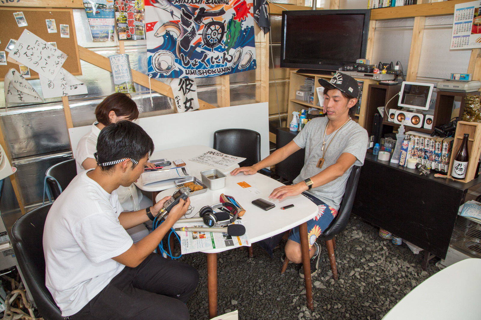 大漁旗やチラシなどがたくさん貼られた壁の前に置かれた白いテーブルの左側に機材を持った男性と女性リポーター、右側に井尻さんが座っている取材の様子の写真