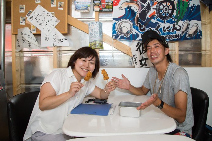 大漁旗やチラシなどがたくさん貼られた壁の前で、串カツを両手に持った女性リポーターと、笑顔の井尻さんが、白いテーブルに向かい合って座っている写真