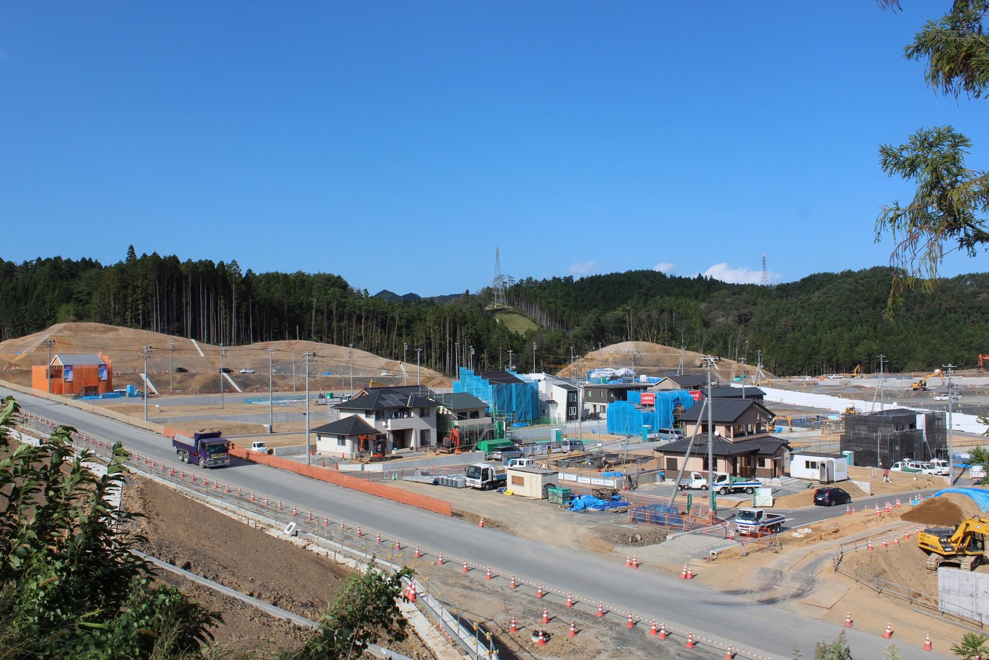南三陸町志津川中央団地の造成地で、整然と並ぶ住宅の建設中の基礎や資材があり、周囲には緑の丘と広い青空が広がる写真