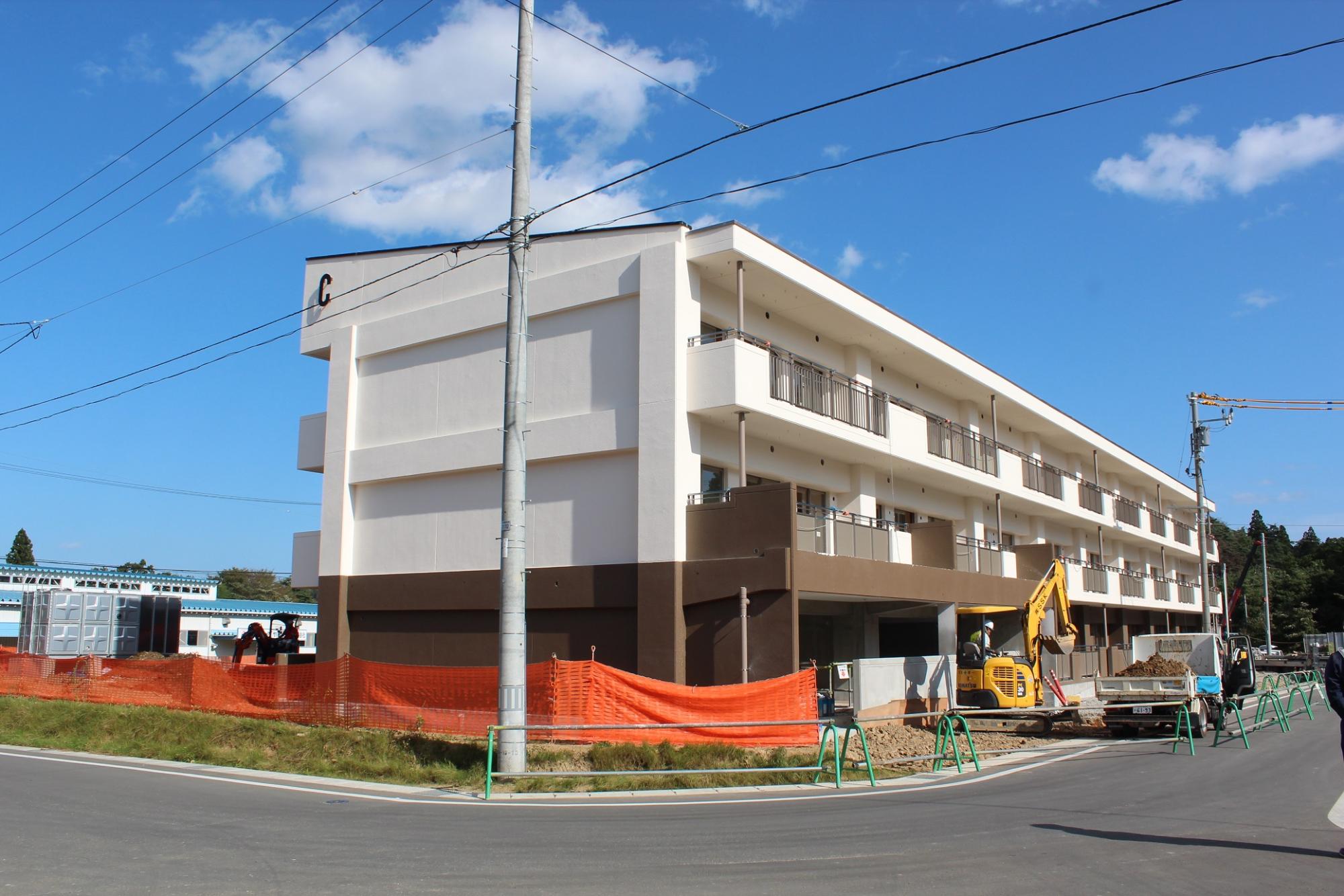青空の下、白と茶色に塗られた3階建ての新築集合住宅の前で重機と作業員が外構工事を行っており、建物の左端には「C棟」と表示された角が見える写真