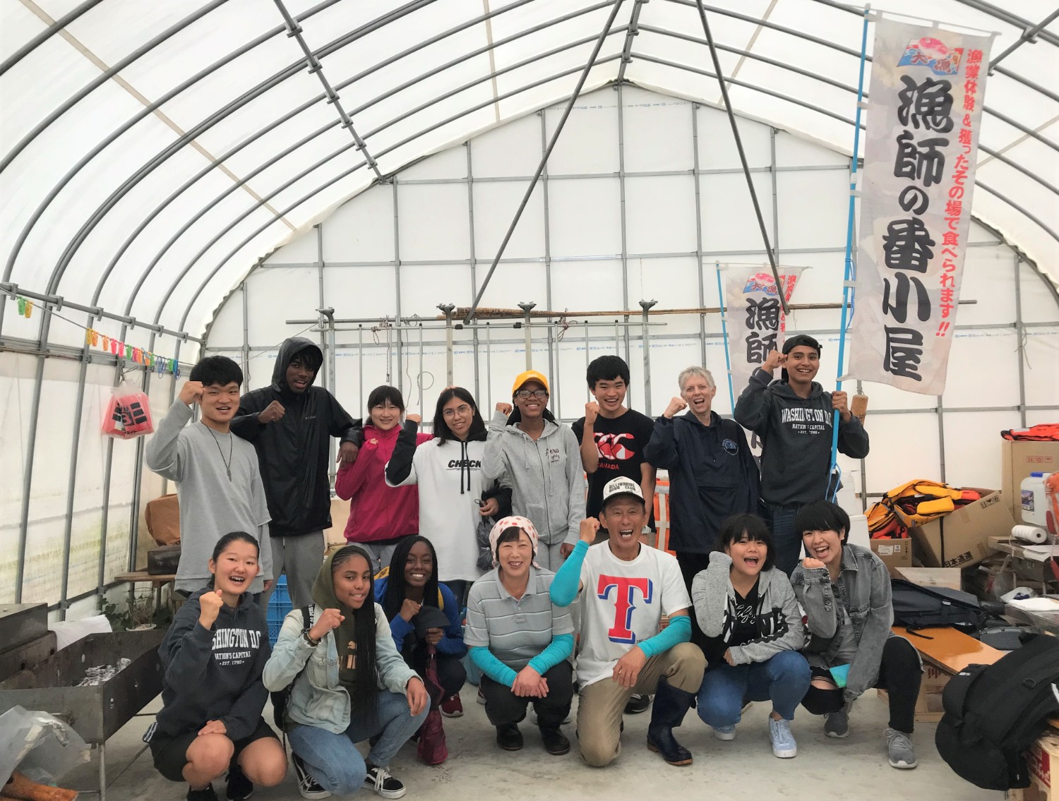 「漁師の番小屋」と書かれたのぼり旗が立っているビニールハウスの中で、日米の高校生たちが地元の人と一緒にポーズを決めて笑顔で2列に並んでいる集合写真