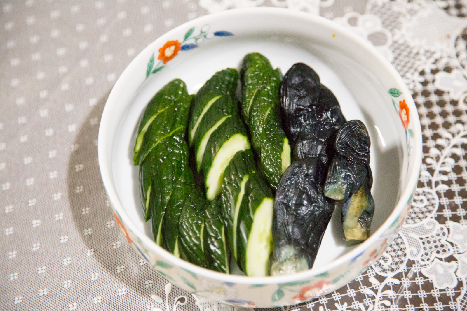 白い器に、緑色のキュウリと黒っぽいナスのぬか漬けが丁寧に盛りつけられ、レース模様のクロスの上に置かれている写真