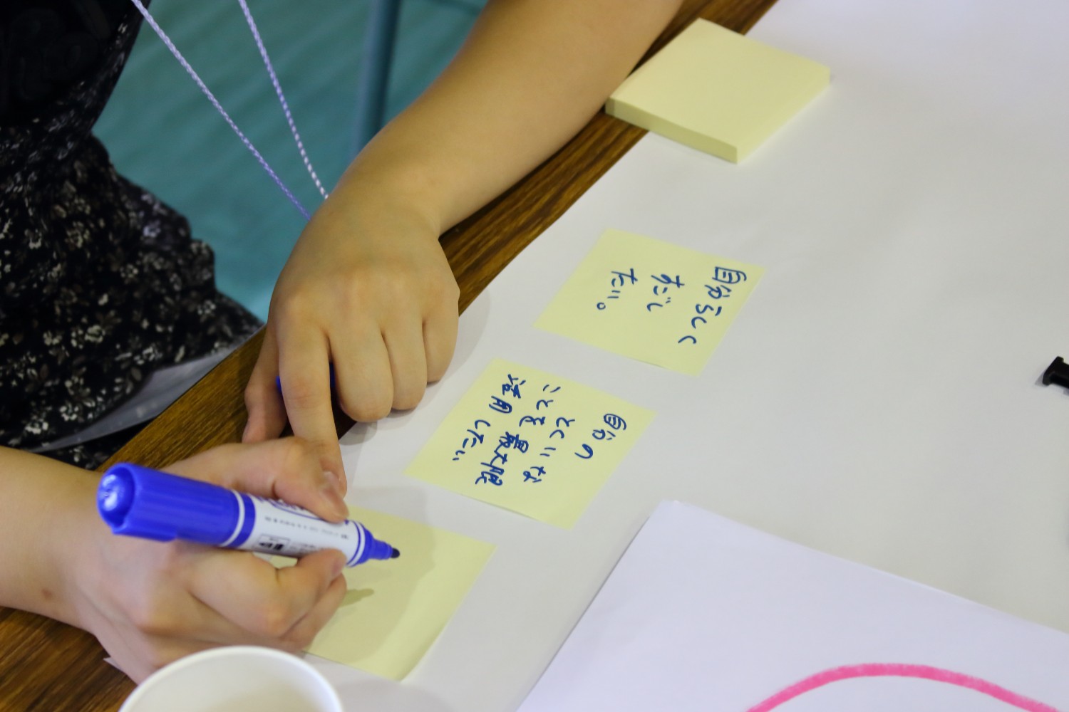テーブルの上で参加者の一人が青いペンで「自分らしくすごしたい」などの文字が書かれた四角形の黄色の付箋が並んでいる手元をアップで撮影した写真