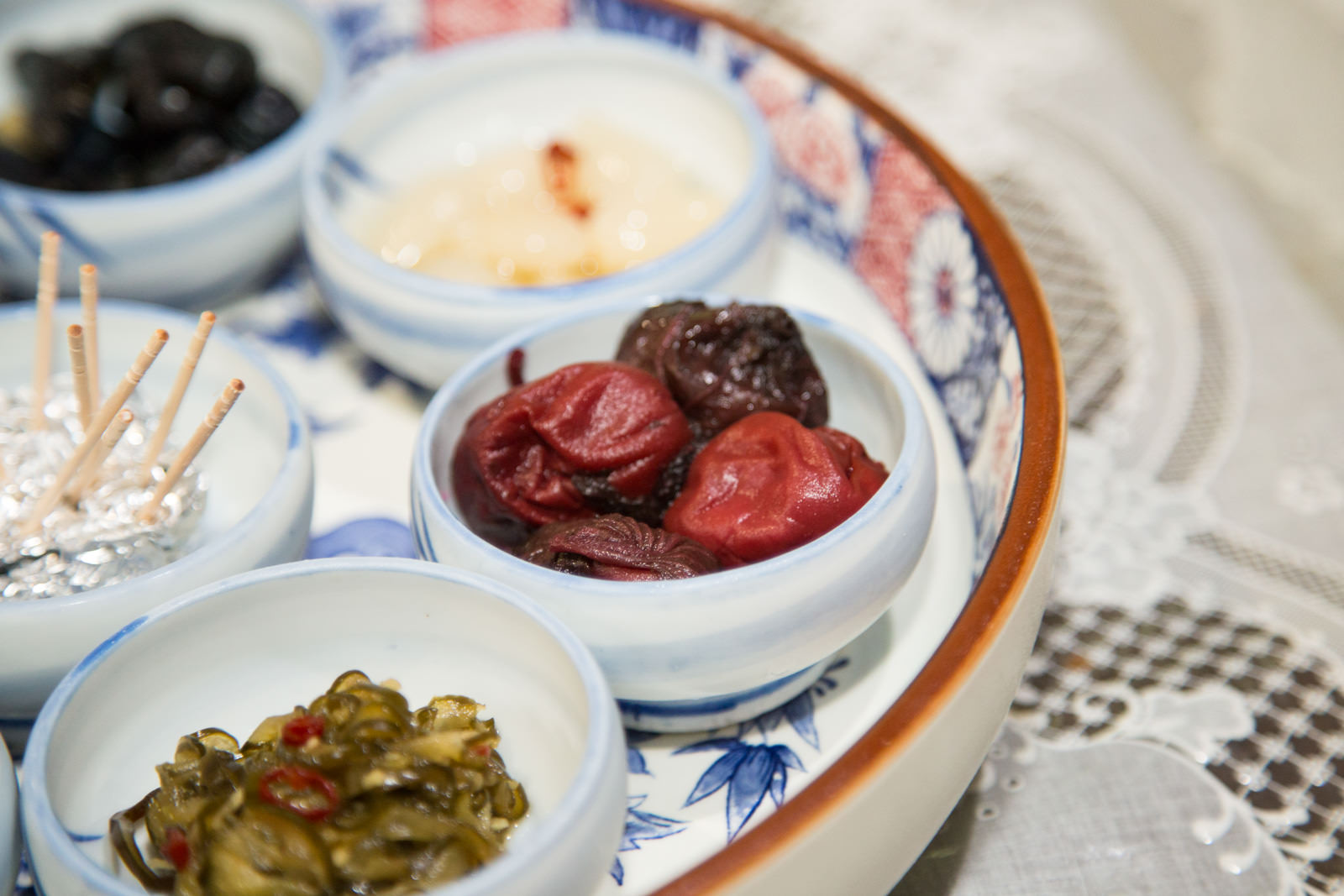 小さな丸い器に赤くしわのある梅干しが入っており、その周りに他の漬物も並んでいる大皿の一部が見える写真