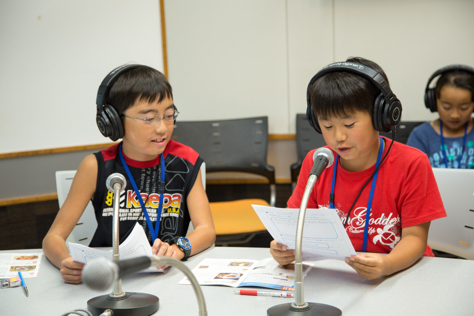 グレーのテーブルに3つのマイクが置かれ、ヘッドホンを着けて座った男子児童2人のうち、1人が手に持った原稿を読んでいる写真
