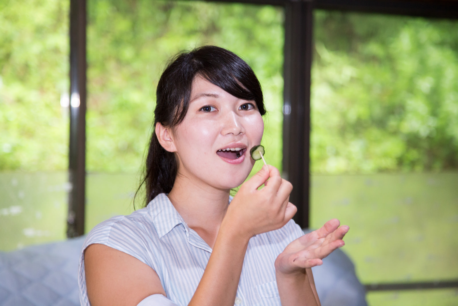 ガラス張りの室内で、女性が笑顔で爪楊枝でさしたキュウリの漬物を手に持ち、口に運ぼうとしている瞬間を写した写真