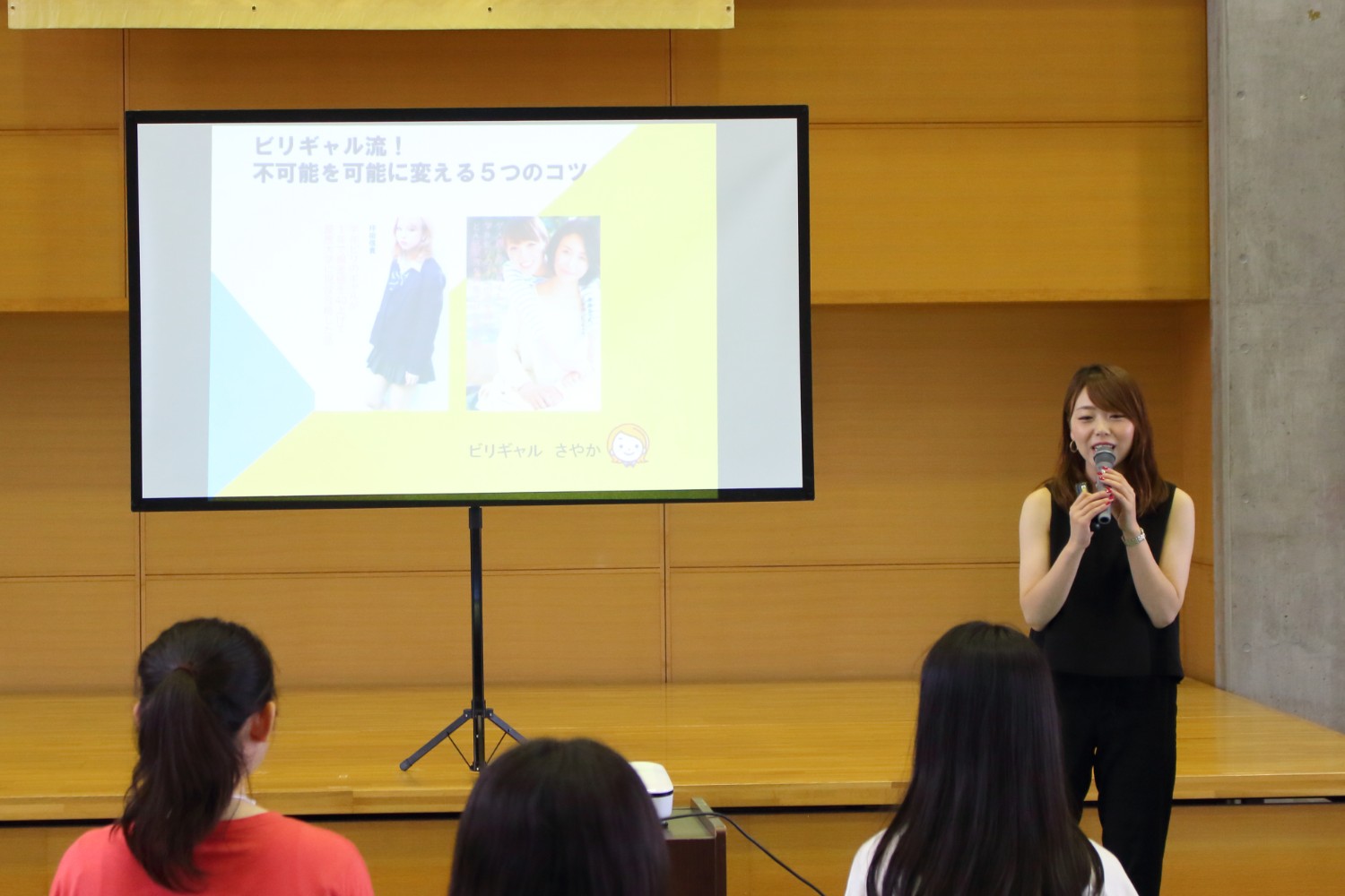 ステージに「ビリギャル流！不可能を可能に変える5つのコツ」と表示されたスクリーンが設置され、壇上で黒い服を着た小林さんがマイクを持って話をしている写真