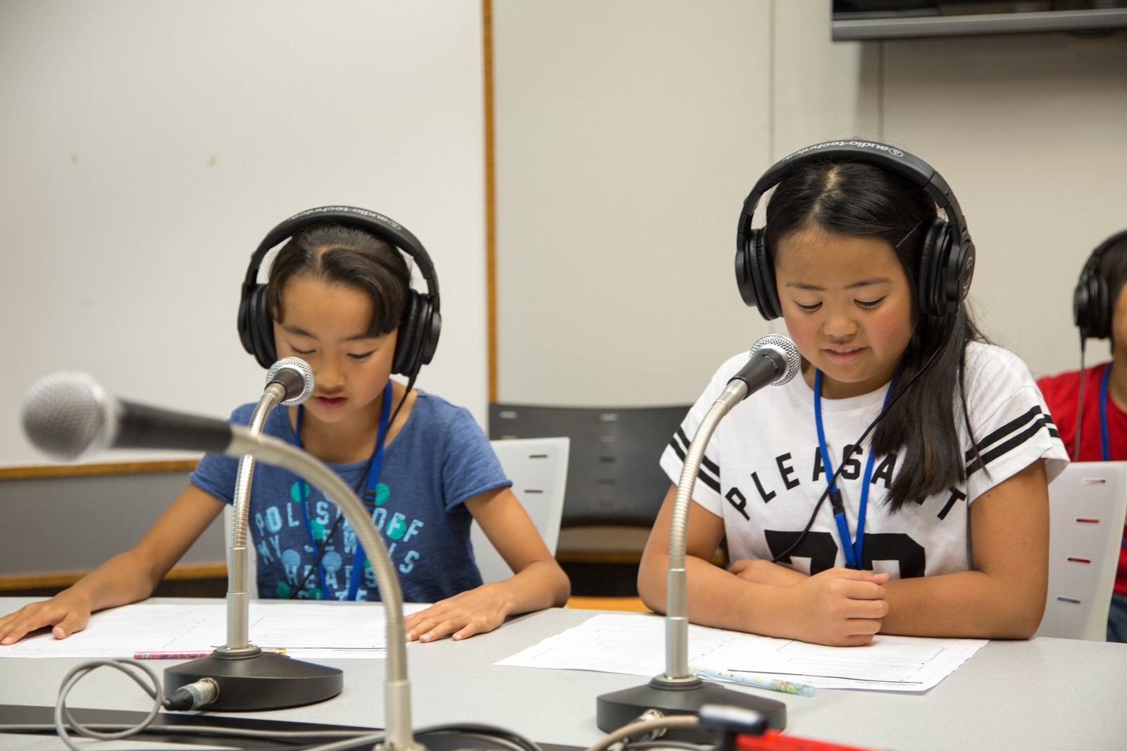 グレーのテーブルに3つのマイクが置かれ、ヘッドホンを着けて座った女子児童2人が、テーブルに置いた資料に目をやっている写真