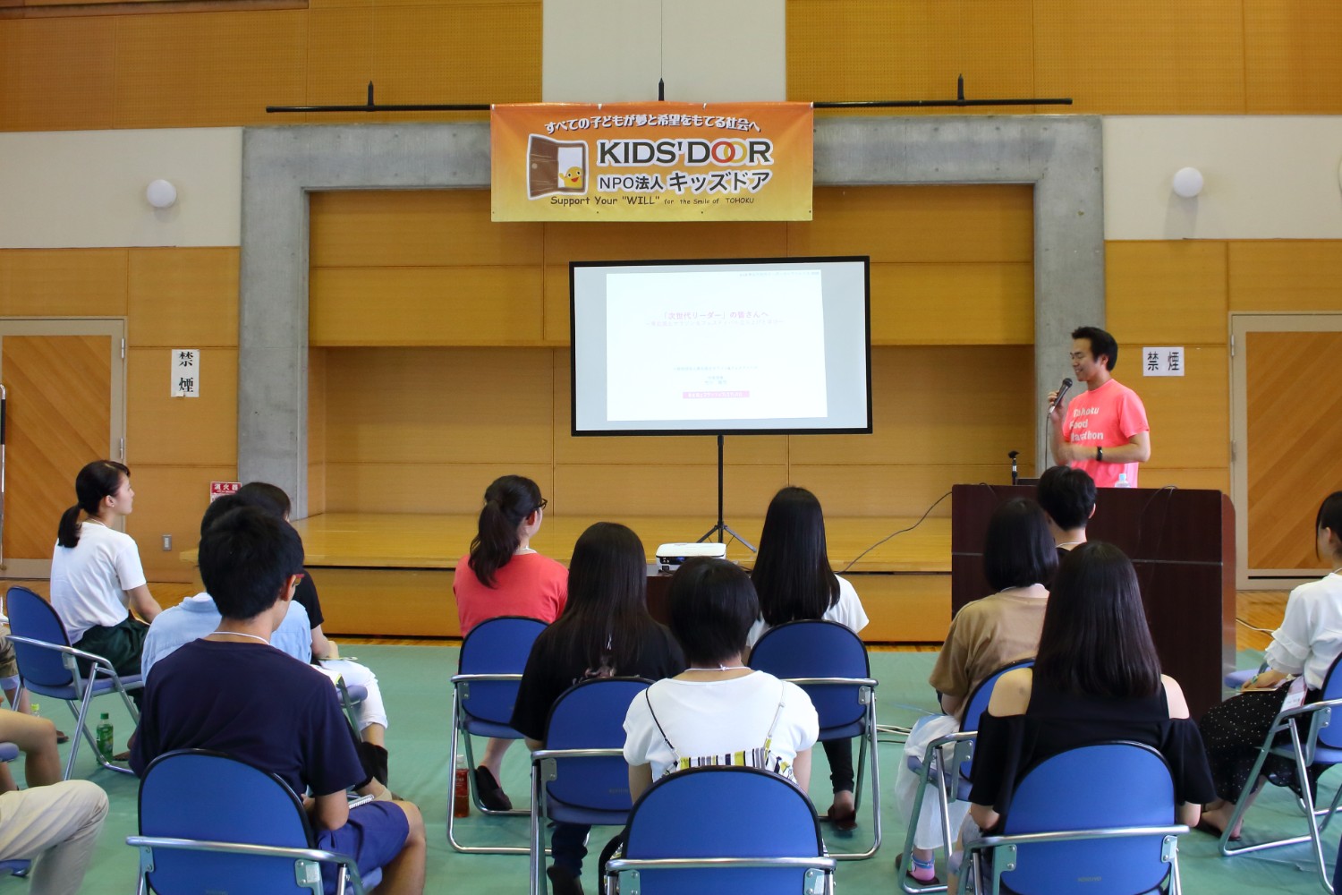 スクリーンを使って資料を見せながらピンクのTシャツを着た男性が壇上でマイクを持って話しており、青い椅子に座った十数人の参加者が真剣に聞いている様子の写真