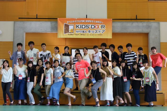体育館のステージ上に「すべての子どもが夢と意欲をもてる社会へ」と「NPO法人キッズドア」のロゴが入ったオレンジ色の横断幕が掲げられ、その下で参加した高校生や関係者がポーズをとり笑顔で並んでいる集合写真