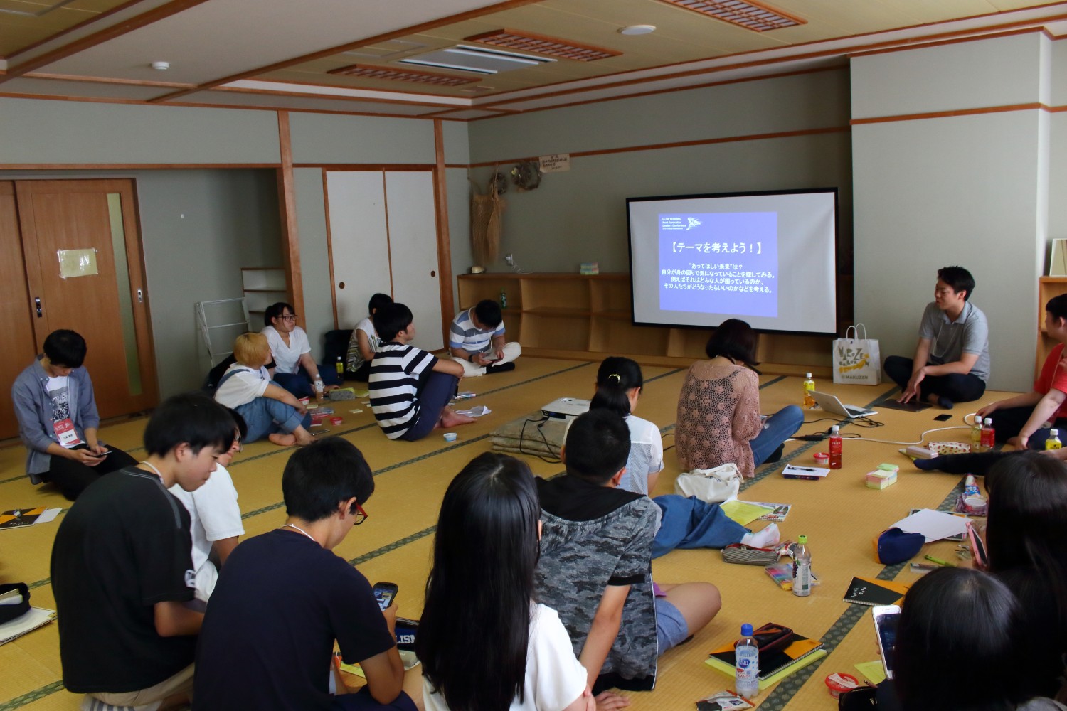畳の部屋で二十人近い若者たちが車座になり、スクリーンに映された「テーマを考えよう！」というスライドを見ている様子の写真