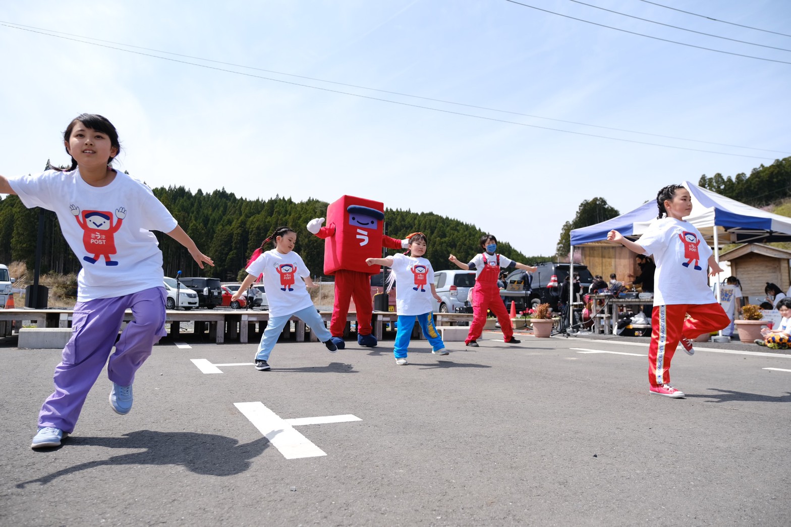 屋外の駐車場のようなスペースで、赤いオーバーオールを着た女性と赤いポストのキャラクターが後ろで一緒に踊って見守るなか、赤いポストが描かれたTシャツを着た子どもたちが両手を広げて元気に飛び跳ねて踊っている写真