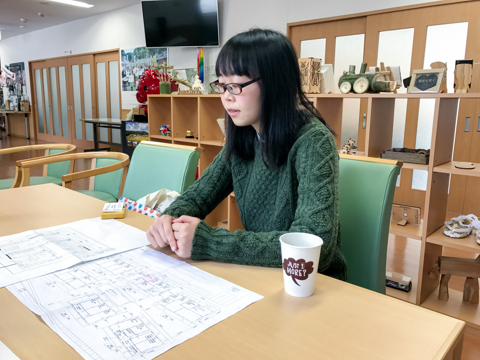 様々な工芸作品が飾られた飾り棚の前で、椅子に座った佐藤さんが、テーブルに広げたレイアウト図に視線を落としている写真