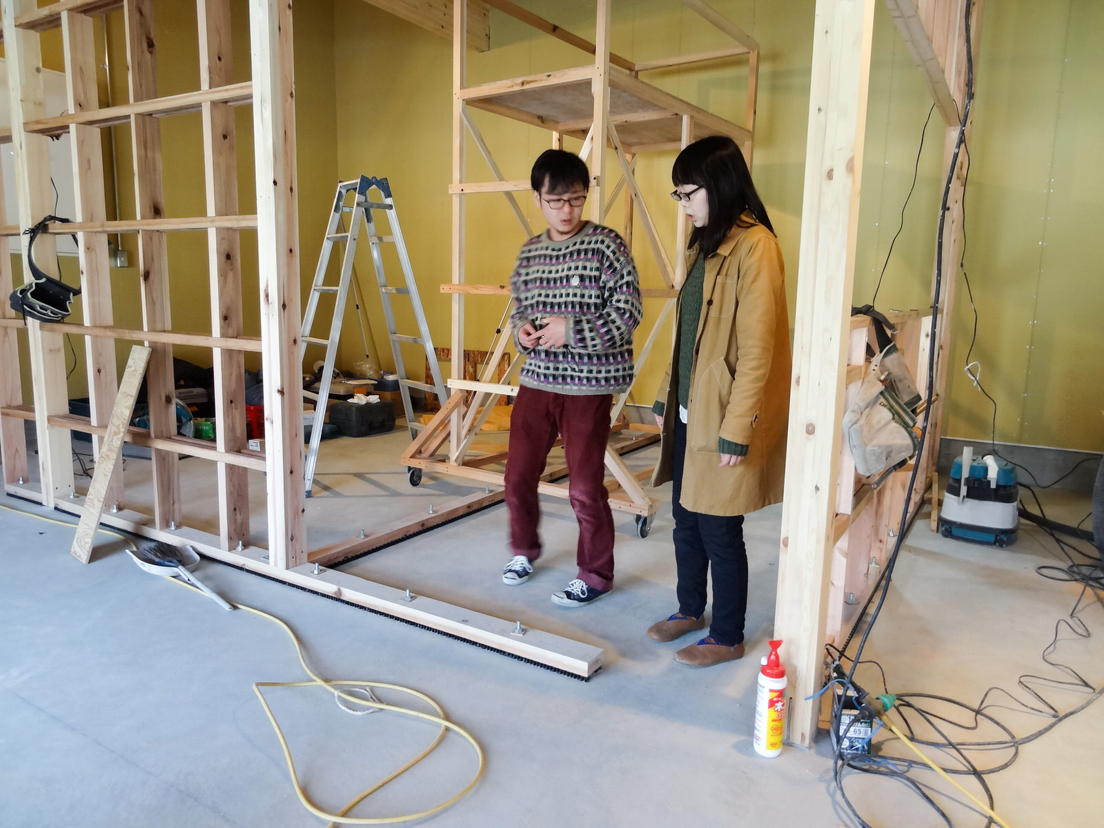 脚立などの道具が置かれ、木の骨組みが見える建設途中の室内で隣同士に立った永井さんと佐藤さんが話をしている写真