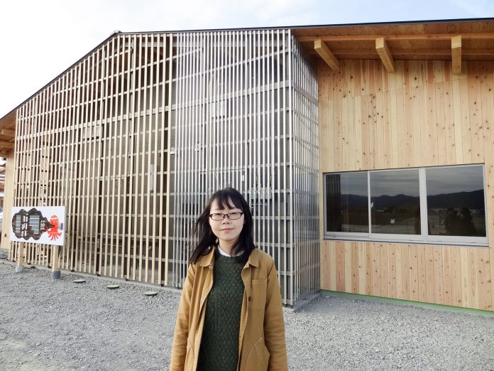 木造で中央あたりが格子状のデザインになった新しい建物の前の砂利のスペースに笑顔で立つ佐藤知里さんの写真