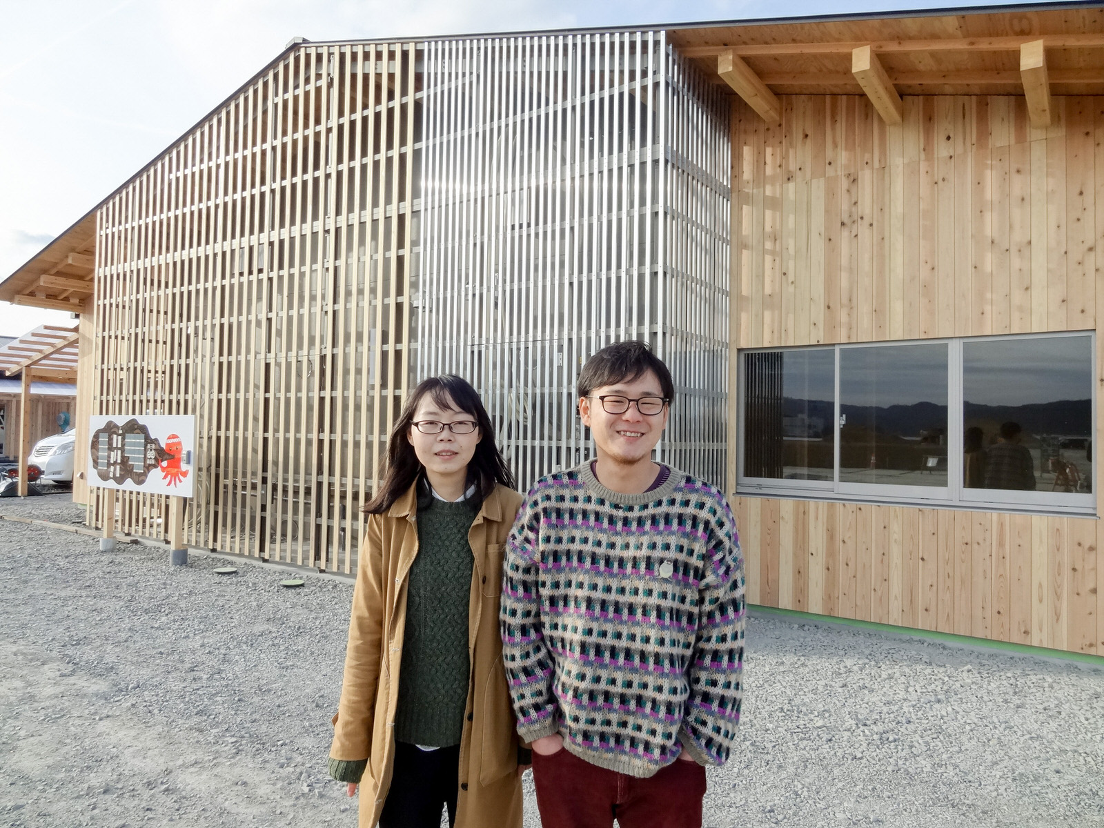 木造で中央あたりが格子状のデザインになった新しい建物の前の砂利のスペースに笑顔で並んで立っている佐藤さんと永井さんの写真
