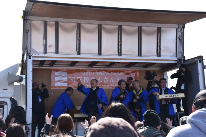 軽トラックの荷台をステージにして、青い法被を着た関係者たちが観客に向けて餅まきをしている様子を写した、南三陸さんさん商店街の開業記念イベントの写真