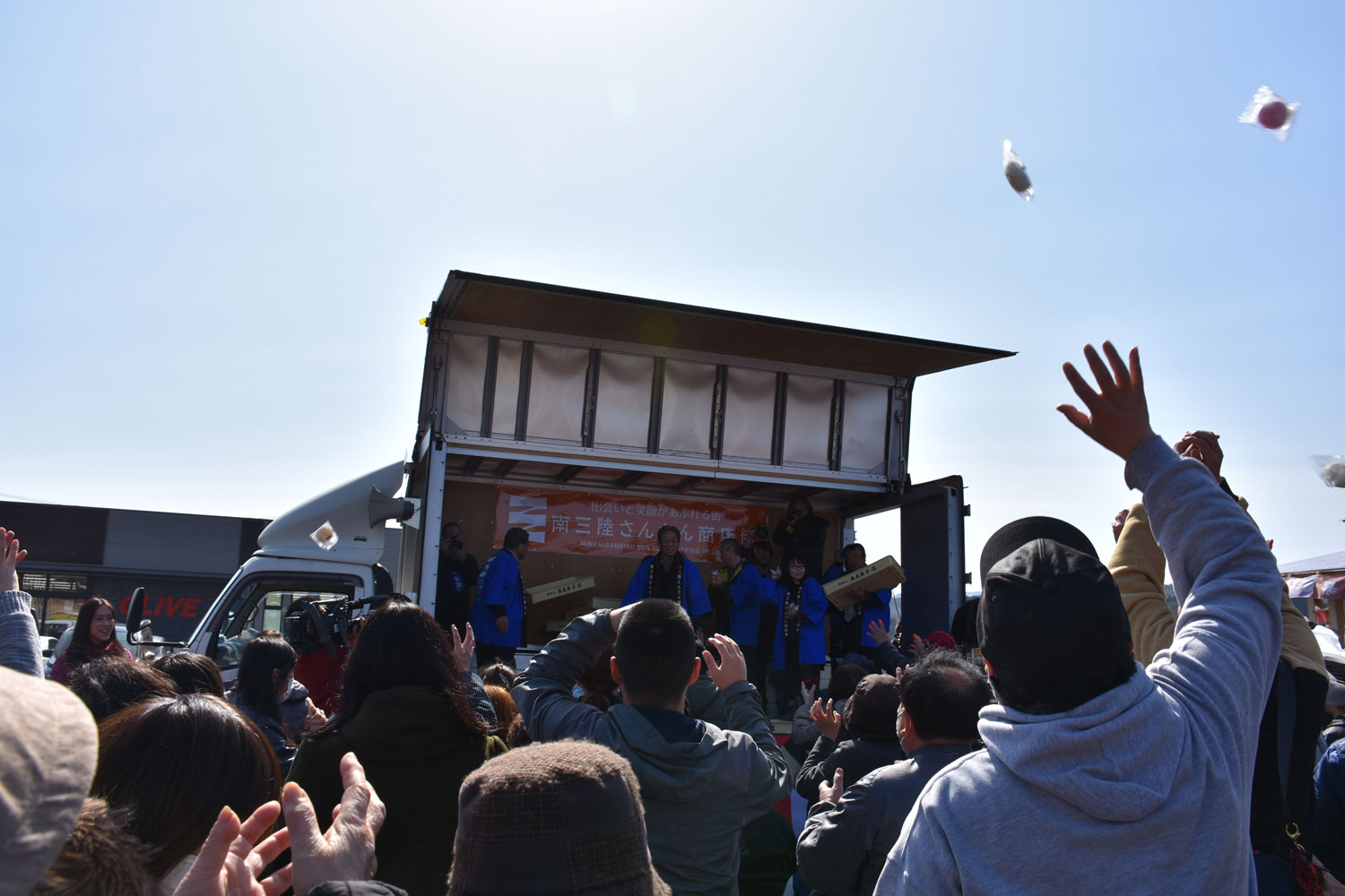 観客たちが手を上げて空中に舞う餅を受け取ろうとしている様子と、ステージ上の主催者たちが餅をまいている様子を写した写真