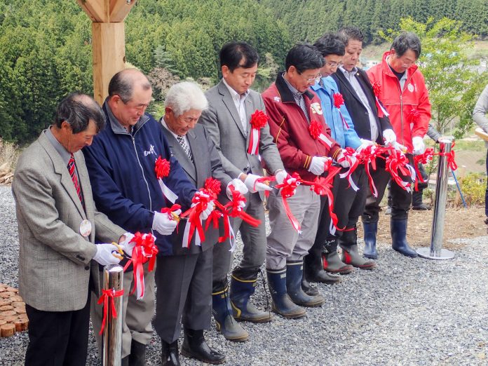 緑の木々を背景に胸に赤い胸章をつけた8人の関係者の男性が横一列に並び、テープカットを行っている様子の写真