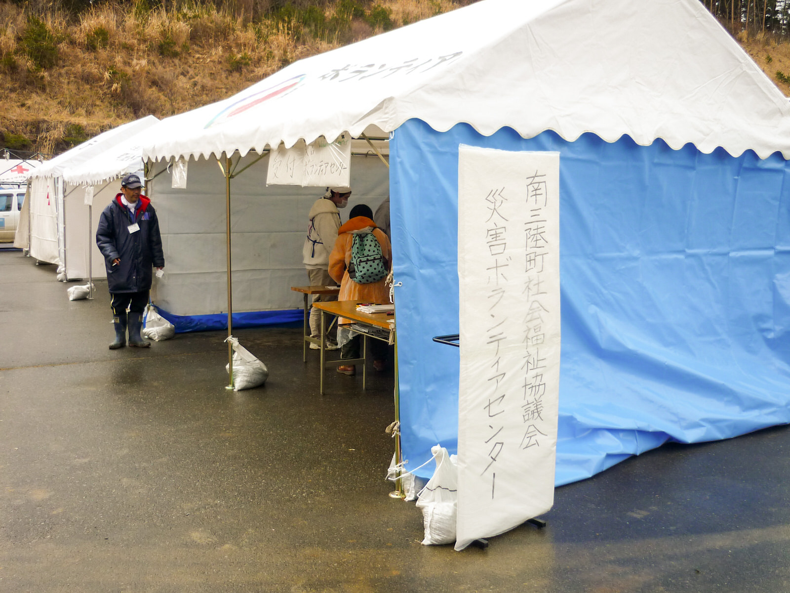 「南三陸町社会福祉協議会 災害ボランティアセンター」と手書きで書かれた看板の横にブルーシートで壁を作った白いテントが立っており、中にテーブルが置かれ人々が活動している様子が見える写真