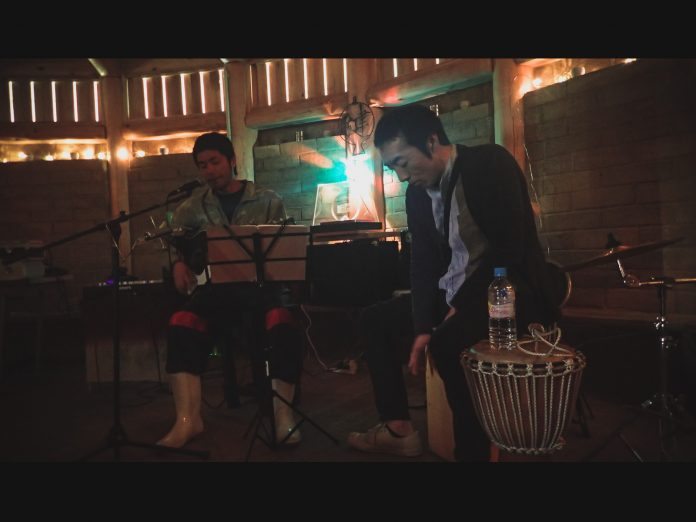 薄暗い木造の屋内で、左側の男性がギターを弾きながら歌い、右側の男性が打楽器のカホンに座って演奏している、静かな雰囲気のライブ演奏の写真