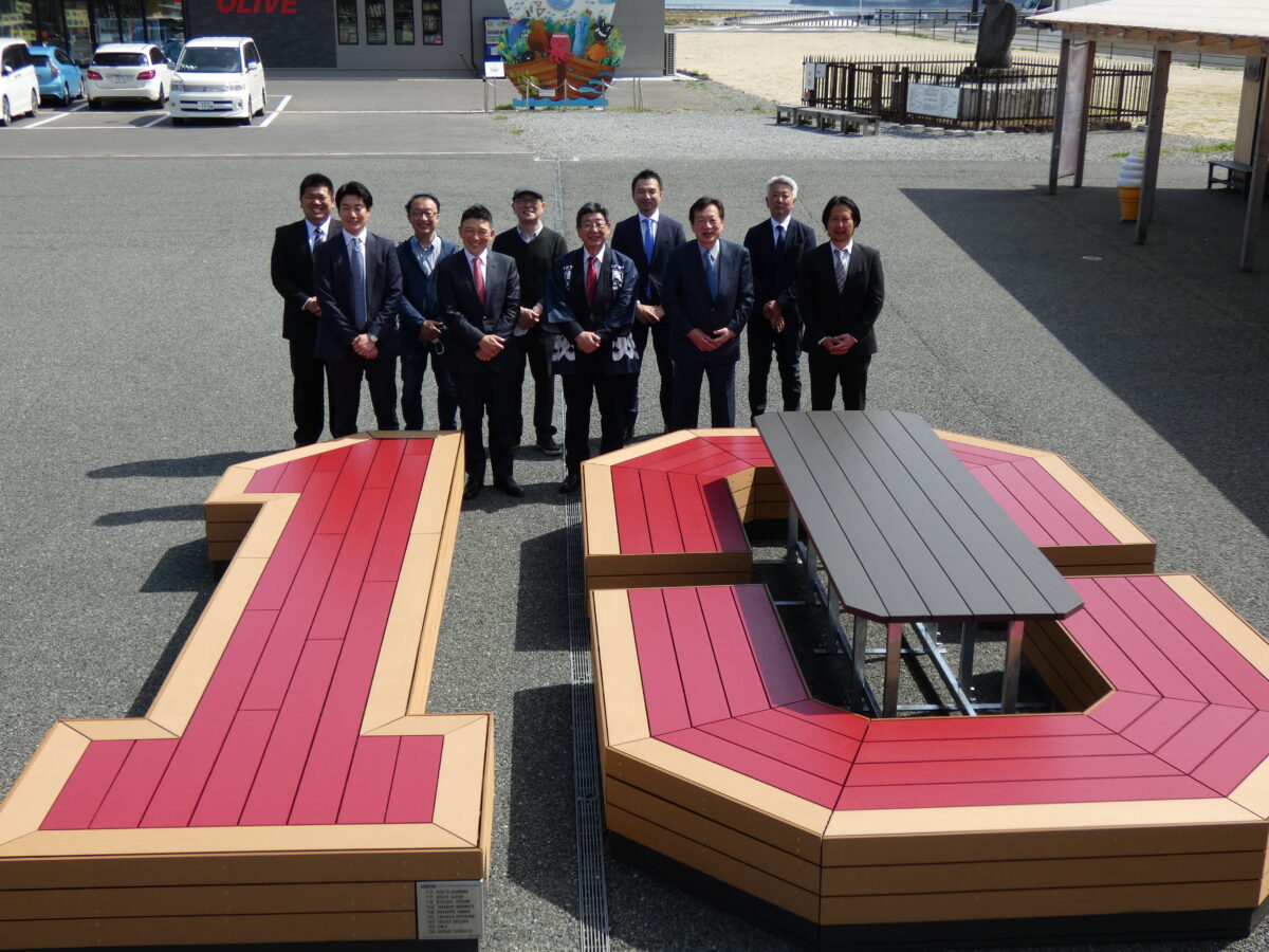赤とベージュでデザインされた巨大な「10」の形のベンチに囲まれたテーブルの後ろに、スーツ姿の関係者たちが整列している、モニュメント前での記念写真