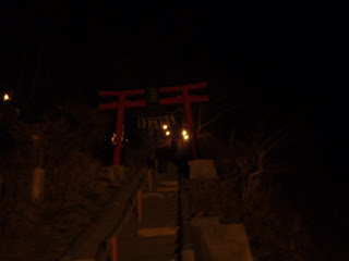 夜の暗闇の中で石段を登る先に赤い鳥居が立ち、その周囲をわずかな灯りが照らしており、階段下から撮影した写真