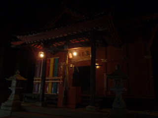 暗闇の中で朱色の拝殿が提灯の明かりにぼんやり照らされ、左右には石灯籠が並び、正面の入口には五色の布がかかっている写真