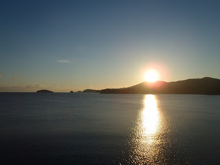 水平線の向こうにある山の稜線から朝日が昇り、穏やかな海面にまっすぐ光の道を作っていて、空は青く澄みわたり、島々の影が浮かんでいる写真