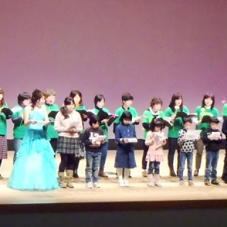 舞台上でドレスを着た女性と緑色のTシャツを着た大人たち、そして前列に並ぶ子どもたちが一緒に歌を歌っている合唱イベントの様子の写真
