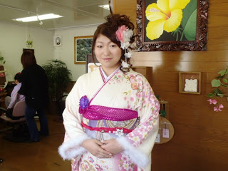 白地にピンク色の花模様があしらわれた艶やかな振袖を身にまとい、上品な髪型と共に笑顔を見せる行場穂乃花（ほのか）さんの写真