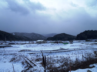 手前には雪に覆われた真っ白な農地が広がり、その奥には連なる山々が遠くに見える冬の風景写真