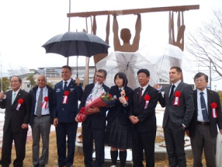 雨の中傘をさしながら復興祈願彫刻「栄光と再生の像」の前で関係者の方々が並んで記念撮影をしている写真