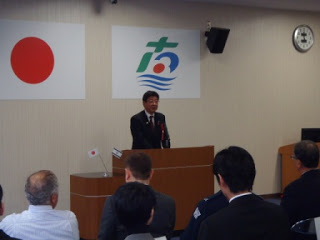 会議室の壇上で佐藤町長がスピーチをしており、背景には日本国旗と南三陸町章が掲げられ、周囲には関係者が椅子に座っている会議室の写真。