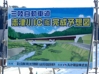 青空と山々が広がる風景の中、緑豊かな川に架かる橋と周辺の道路が描かれ、インターチェンジの構造がわかりやすく示された、三陸自動車道志津川IC（仮）完成予想図の案内版の写真