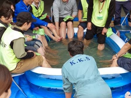 ビニールプールの周りに人々が座り、STAFFと書かれたベストを着た人達が足湯につかっている写真