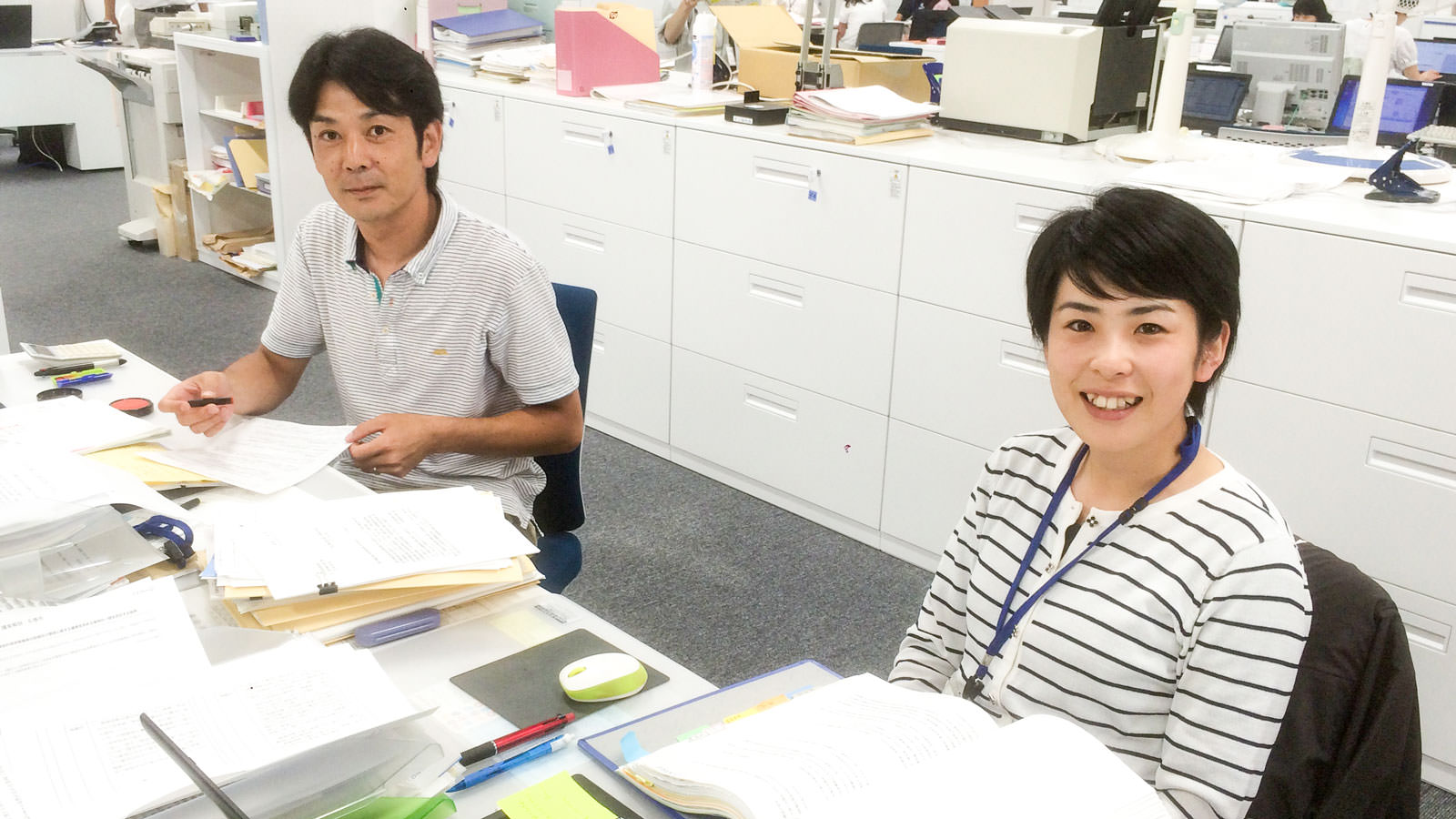 白い書類棚が並ぶ室内で、たくさんの書類が置かれたデスクの席に着いた阿部さんと佐々木さんがカメラを見ている写真