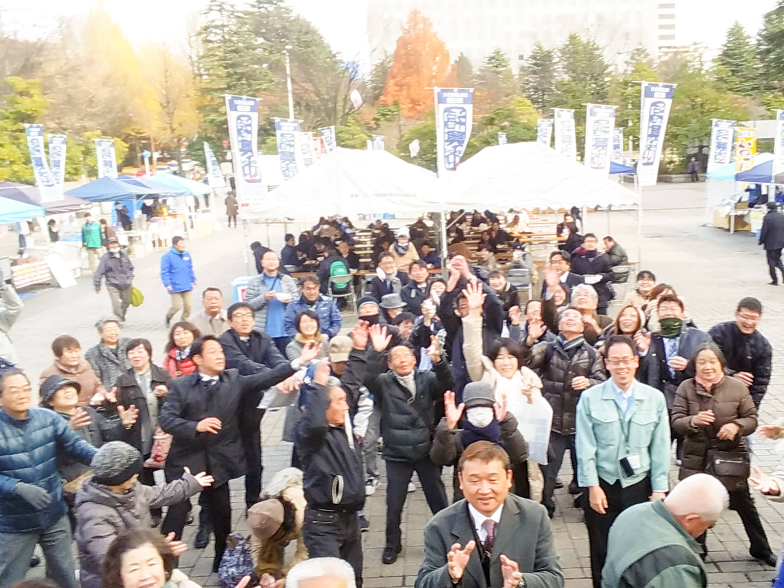 白いテントやのぼり旗が立ち並ぶ福興市の会場で、大勢の人々が空に向かって手を上げ餅を掴もうとしている様子の写真