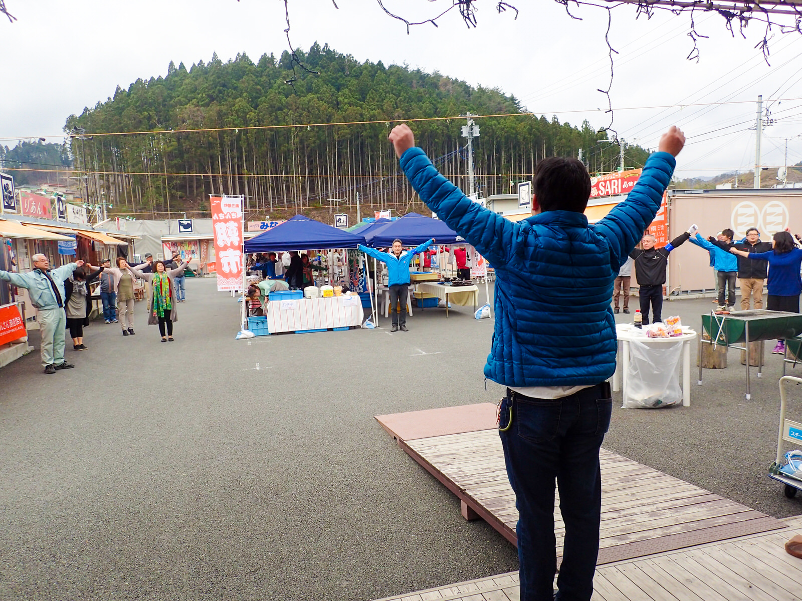ステージの上で両腕を上に伸ばしている本間さんと、ステージ前の店舗やテントの前や通路に立ち、同じ体操のポーズをしている人々の写真
