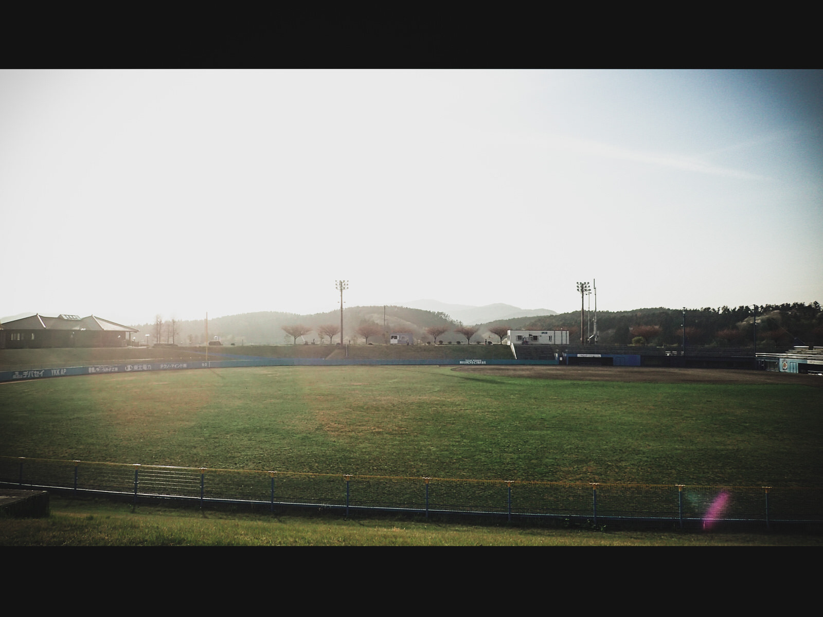 芝生の野球場に夕日が差し込み、奥にはなだらかな山々が見える写真