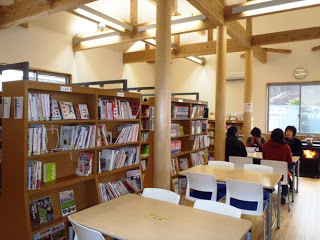 木の柱や梁が見えるあたたかみのある図書館の室内で、左側には本棚が並び雑誌や本が収納され、右側にはテーブルと椅子が配置されて利用者が座っており、奥には大きな窓から外の景色が少し見える室内写真