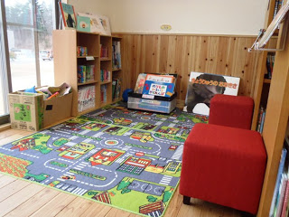 カラフルな道路と建物が描かれたマットが敷かれ、赤い椅子、本棚、窓際に段ボール箱が置かれている子どもスペースの写真。