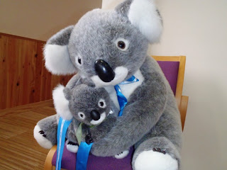 大きなぬいぐるみのコアラが小さなぬいぐるみのコアラを抱いて木製の椅子に座っている写真