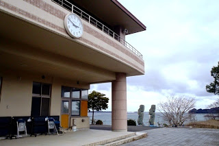 海に面した建物の玄関先に時計が設置されており、奥には波穏やかな海とモアイの石像2体が見える写真