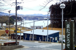 「またどうぞ」と書かれた柱の向こうに、坂道と青い建物、その先に海が見える写真