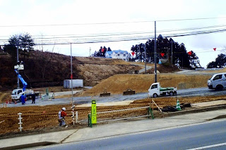 数台の作業車両と作業員が整地作業を行っており、奥には丘の上の建物が見える写真